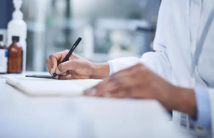 A stock image showing a black woman scientist's hands making notes in a laboratory.