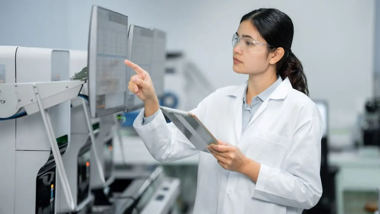 A female scientist with brown hair in a white lab coat pointing at a mounted monitor while holding an iPad